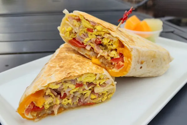 Relish a hearty wrap featuring fluffy scrambled eggs, savory grilled smoked ham, caramelized red onions, vibrant red bell peppers, melted yellow cheddar cheese, crispy hash browns, house-made garlic aioli, and a touch of maple syrup, all wrapped in a warm flour tortilla.
